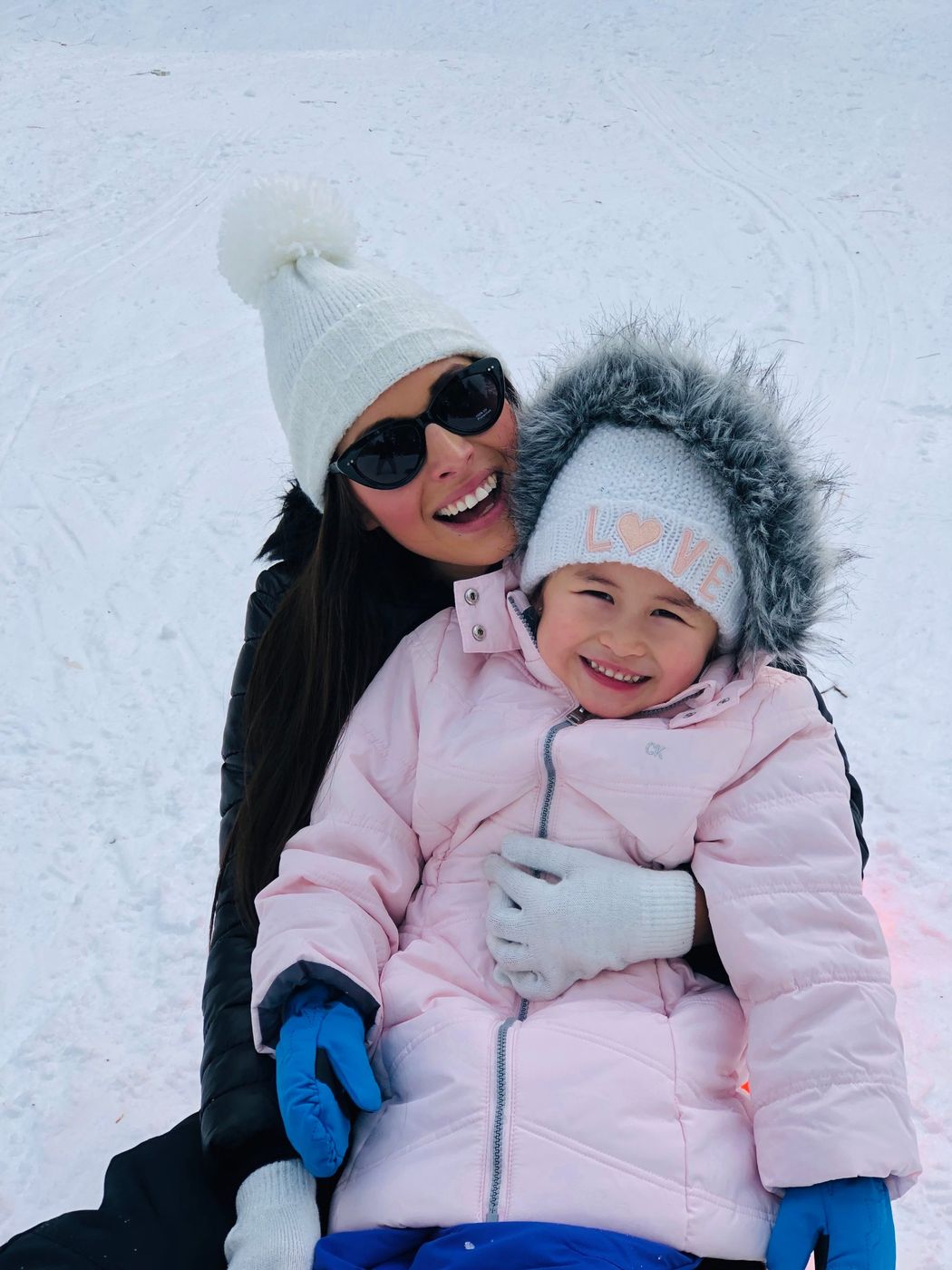 A smiling woman and young girl bundled up in winter hats and coats, sitting in the snow