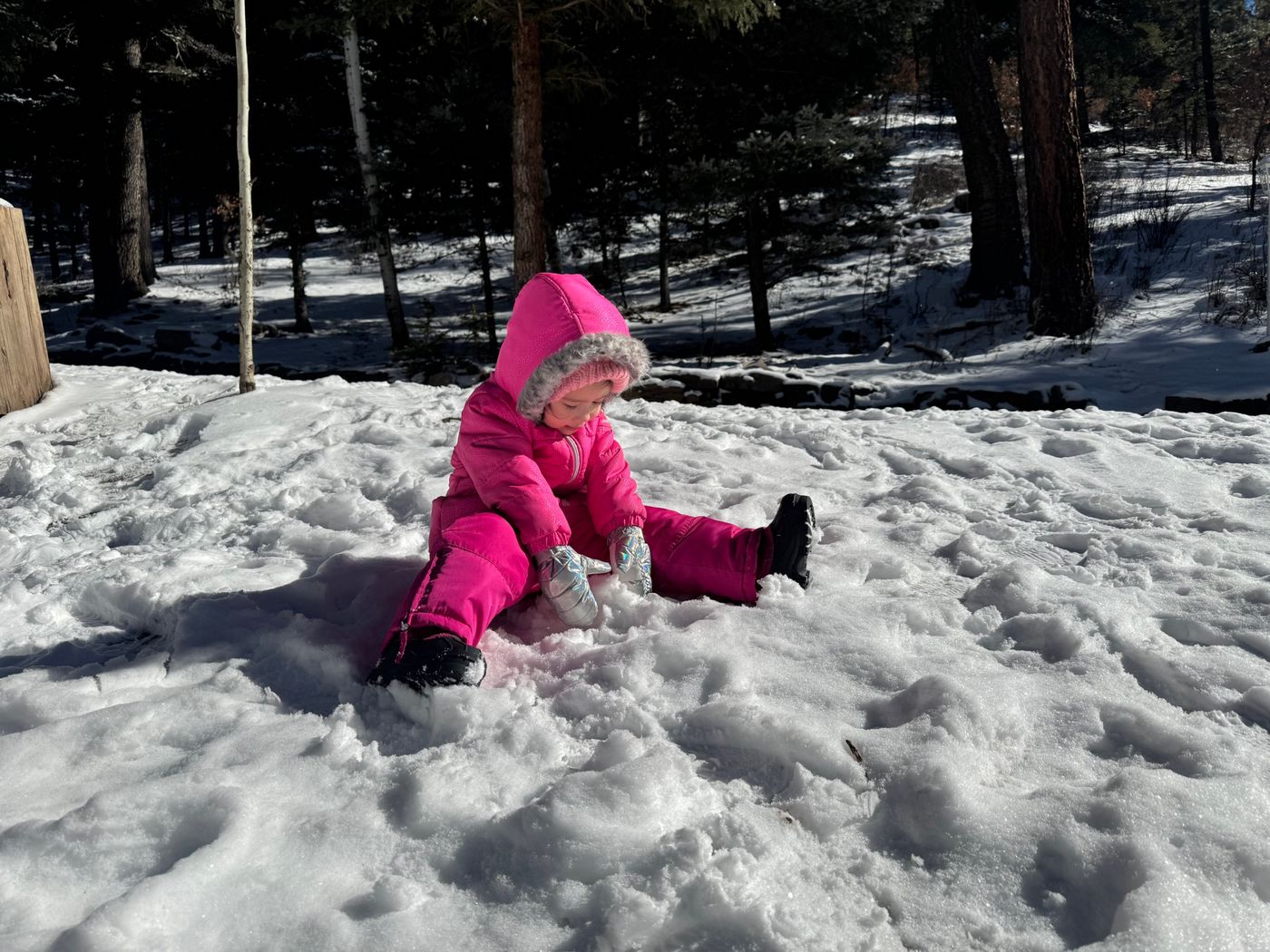 A toddler in a pink snowsuit sits in the snow, playing with it